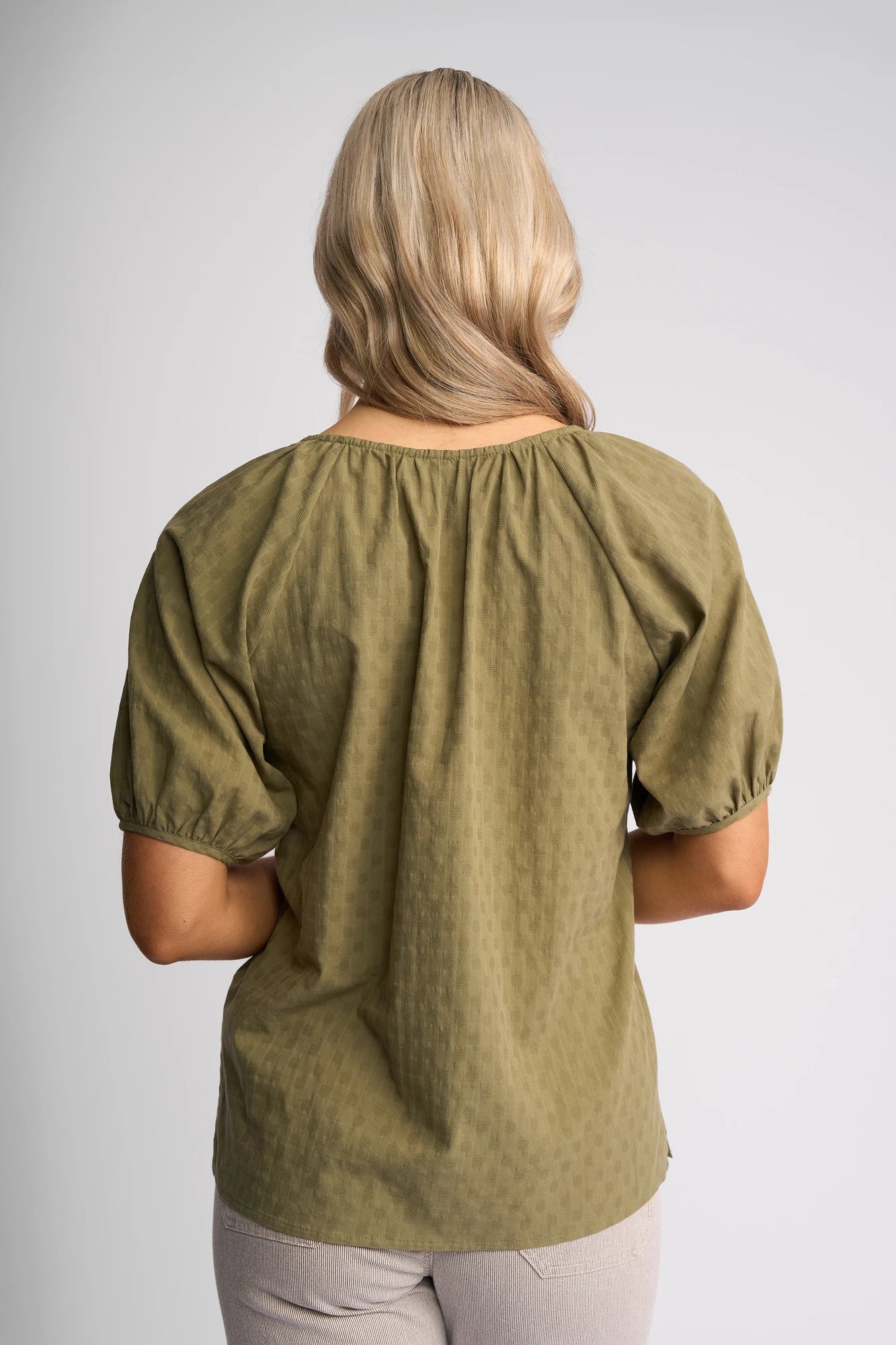 A person with long, wavy blonde hair is shown from behind, wearing a loose-fitting Vassalli Pitch Top in khaki—a comfortable cotton top—paired with light-colored pants against a plain background.