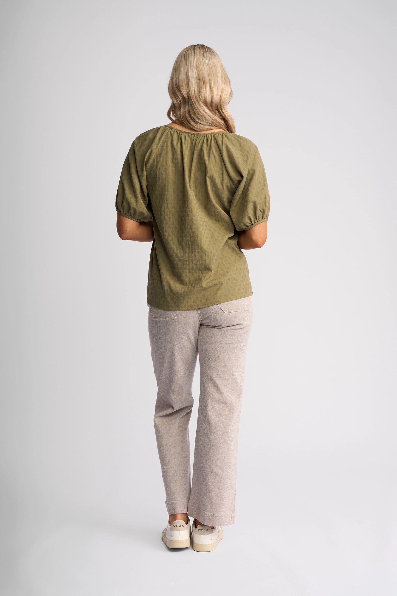 A person with long blonde hair stands facing away, wearing a Vassalli Pitch Top in Khaki, paired with beige pants and white sneakers, against a plain light gray background.