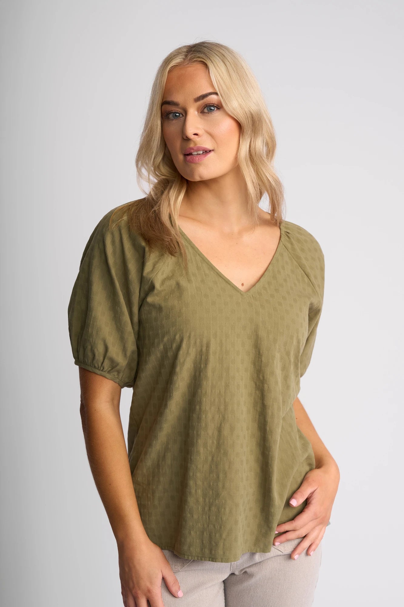 A woman with blonde hair wears an olive green (Khaki) Vassalli Pitch Top—a stylish cotton top featuring a V-neck and short sleeves—and light pants, standing against a plain light gray background while looking slightly to the side.