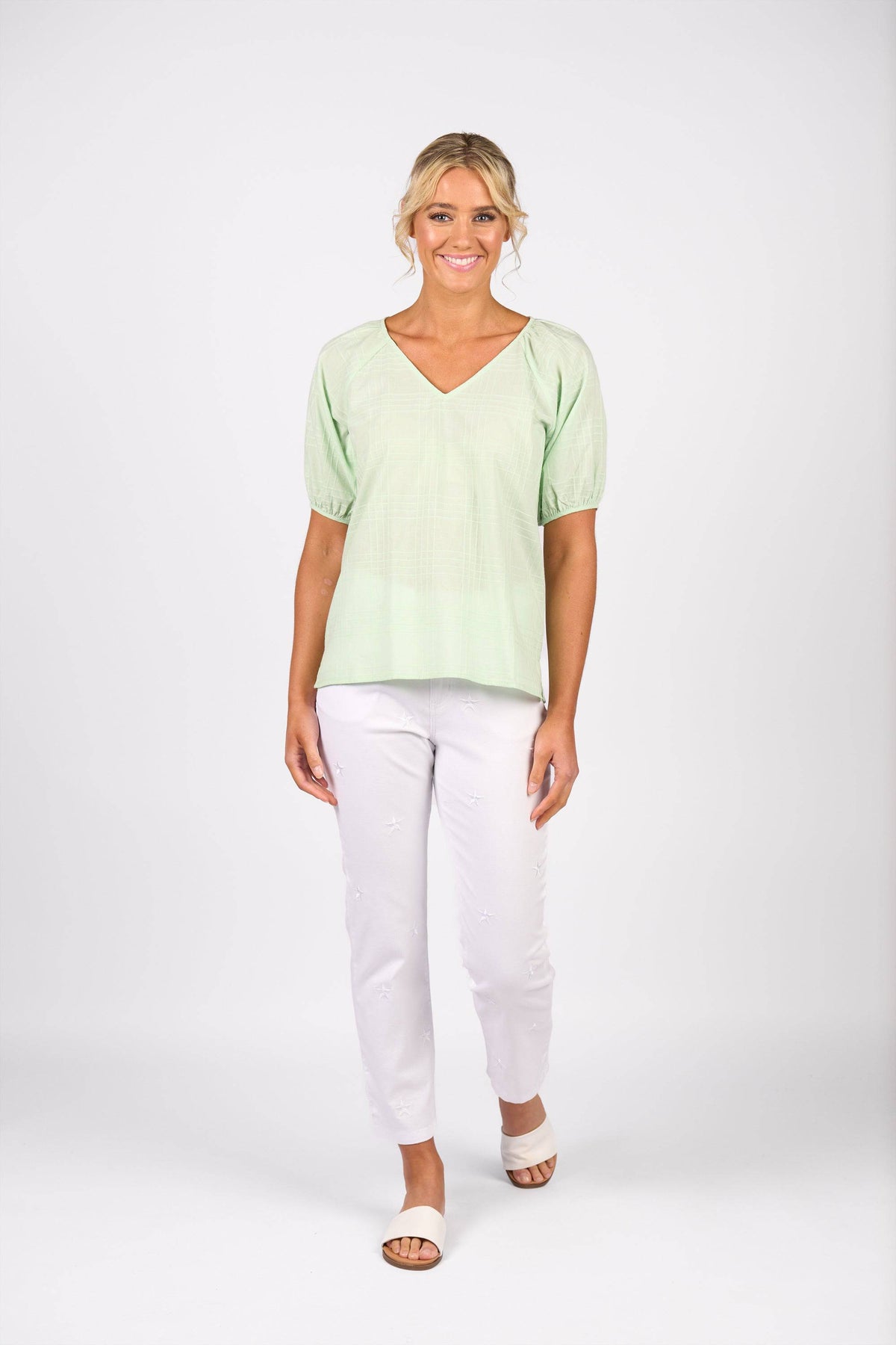 A woman stands smiling against a white background, wearing the Vassalli Pitch Top in mint, styled with white pants and beige sandals. Her hair is in a loose updo.