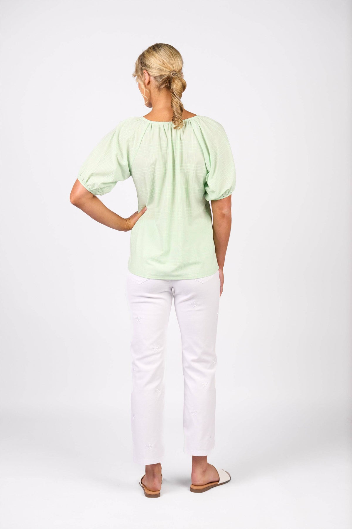 A woman with blonde hair in a braid stands facing away, wearing the Vassalli Pitch Top - Mint, white pants, and white slip-on shoes against a plain white background.