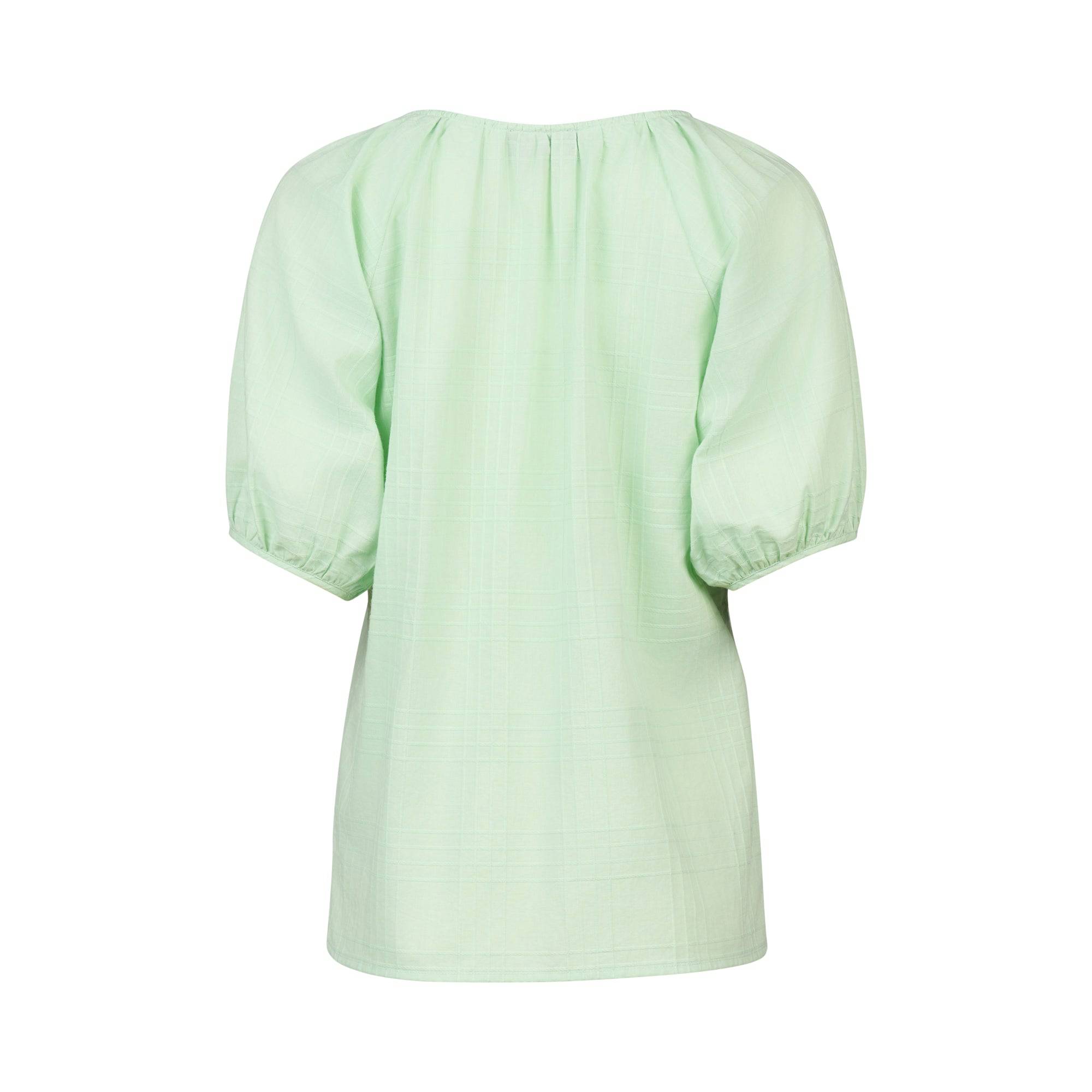The Pitch Top - Mint by Vassalli is a light green cotton blouse with short puff sleeves, a loose fit, gathered neckline, and airy fabric. Shown from the back, it features a simple design with no visible buttons or embellishments.
