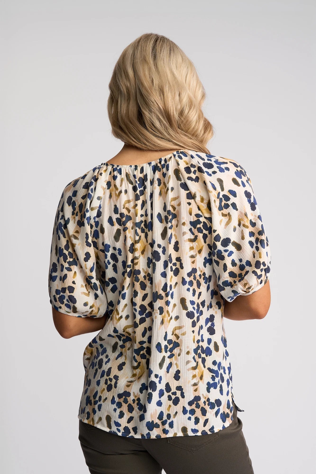 A woman with wavy blonde hair, seen from behind, wears the Vassalli Pitch Top - Leo—a lightweight short-sleeve V-neck top featuring a striking blue, yellow, and black abstract print—paired with dark pants against a plain light background.