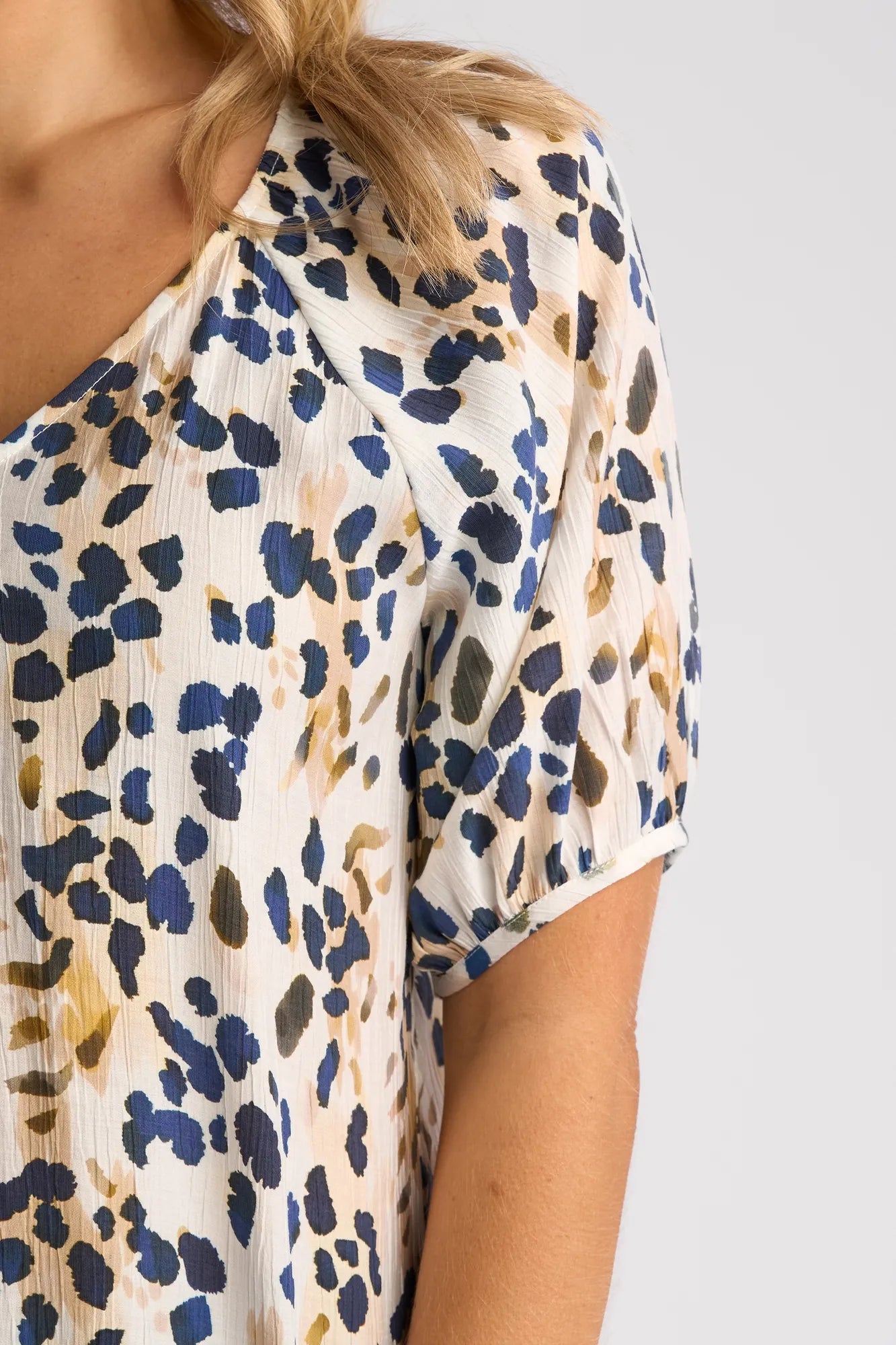 A close-up of a woman’s upper torso wearing the Vassalli Pitch Top - Leo, a short-sleeved, lightweight blouse with a blue, black, and beige abstract print. The top features a V-neckline, and her blonde hair rests partially on her shoulder.