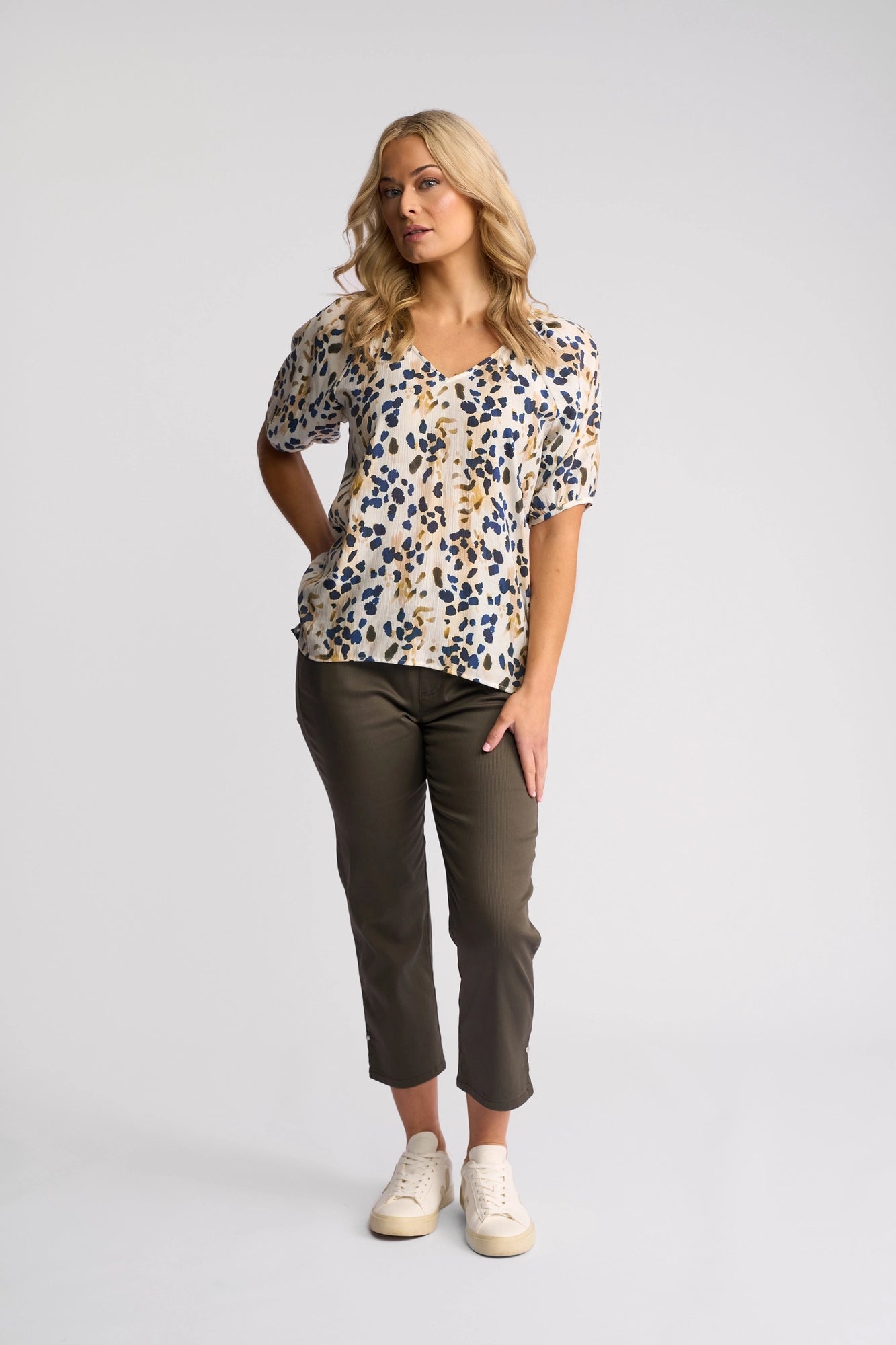A woman with blonde hair stands in a studio with a neutral background wearing the Vassalli Pitch Top - Leo, featuring short sleeves, an abstract print, and a V-neckline, paired with olive green pants and white sneakers.