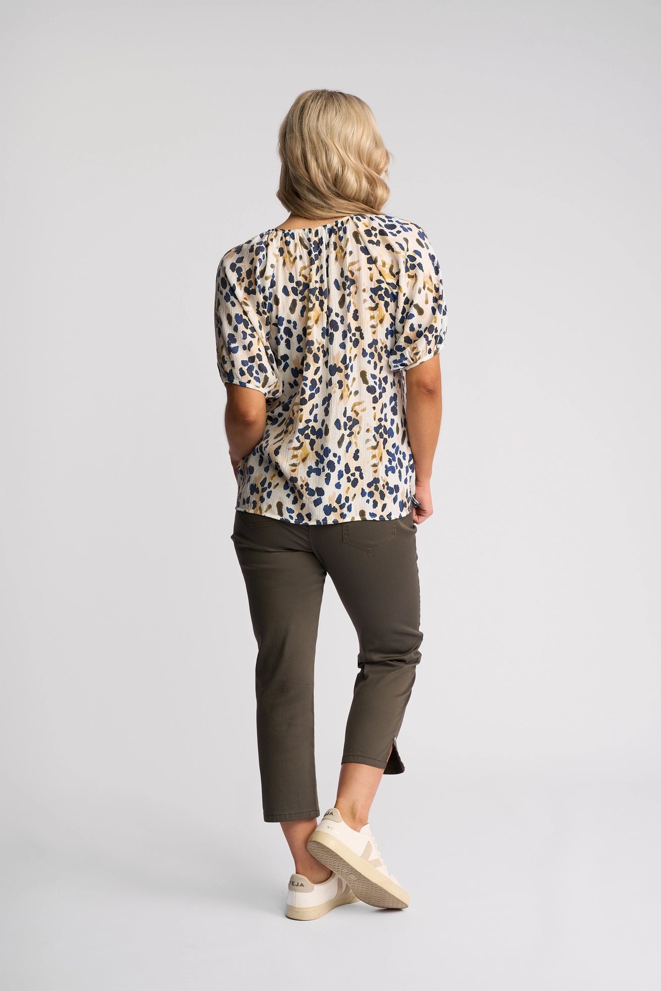 A person with blonde hair, wearing the Vassalli Pitch Top - Leo—a lightweight, abstract print short-sleeve blouse with a V-neckline—paired with olive-green cropped pants and beige sneakers, stands facing away against a plain light background.