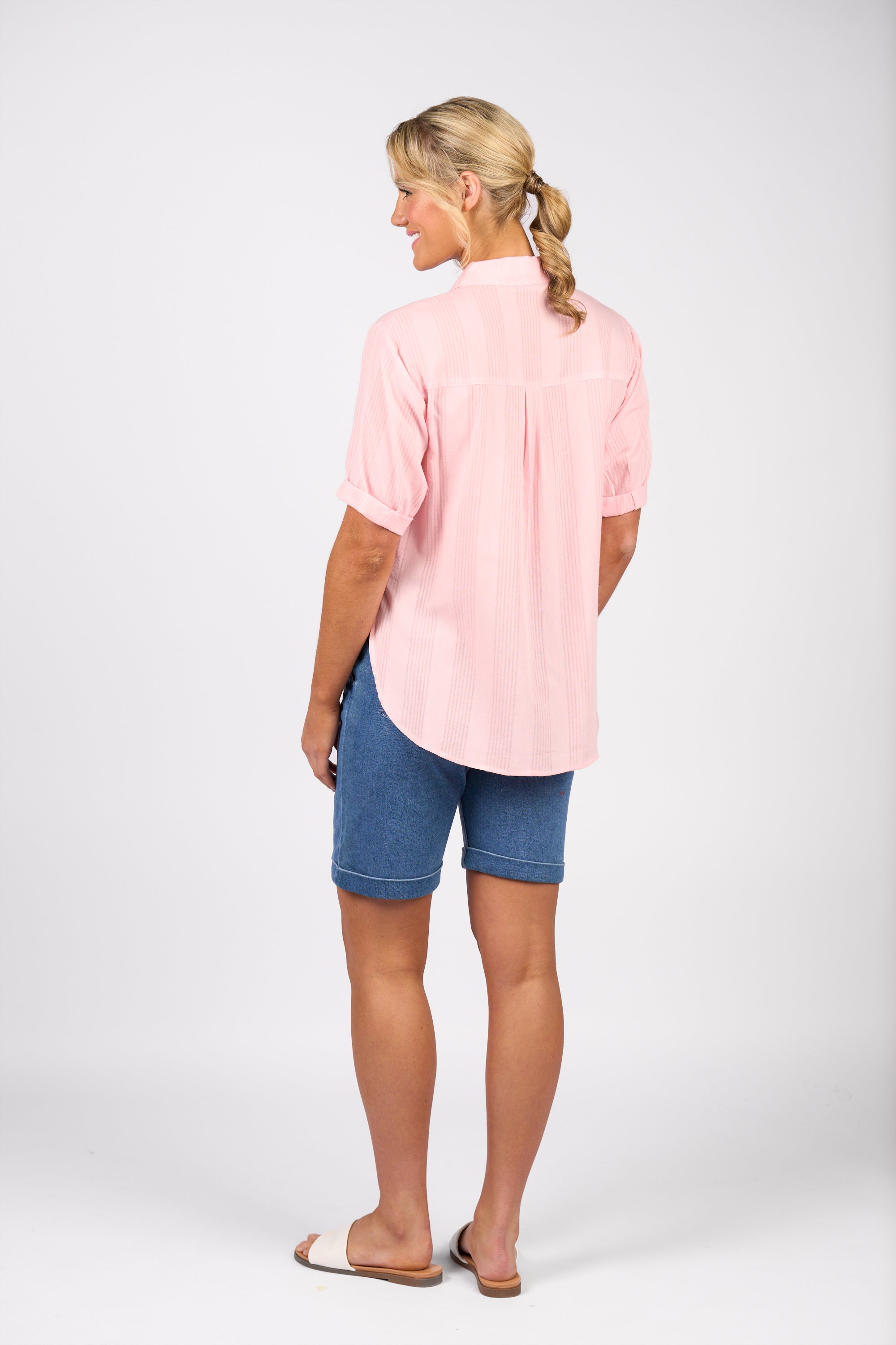 A person with blonde hair in a low ponytail faces away, wearing the Vassalli Shirt - Pink Stripe, blue knee-length shorts, and sandals against a plain white background.