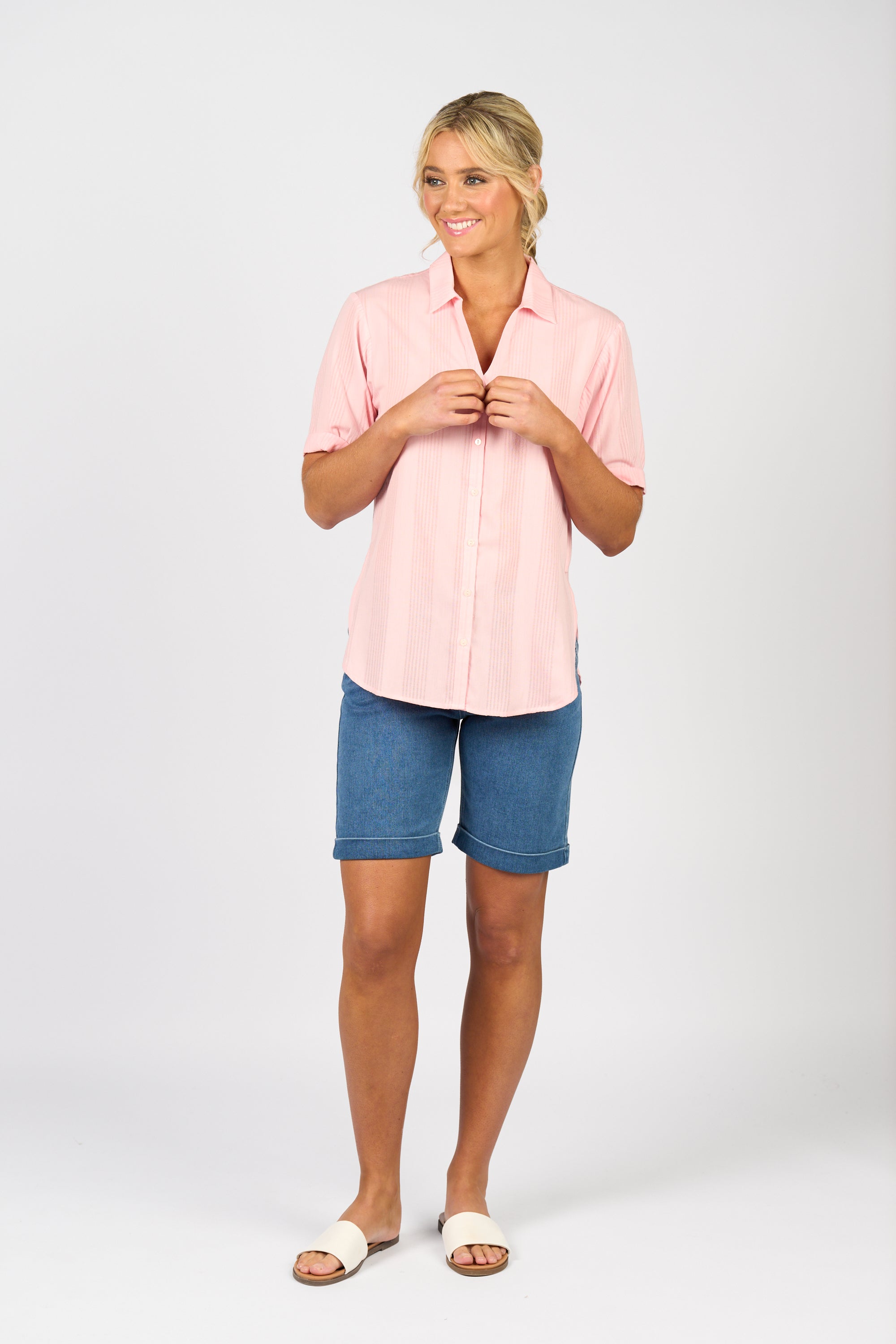 A smiling person stands against a plain background, wearing the Vassalli Shirt - Pink Stripe, blue knee-length denim shorts, and white slide sandals. Their hands are lightly touching the shirt near the collar.