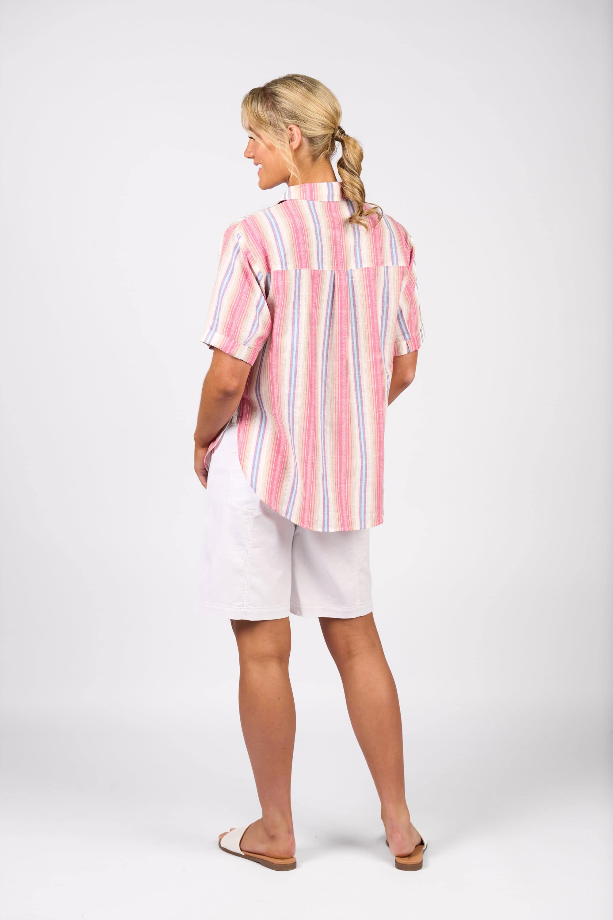 A woman with blonde hair in a ponytail stands against a plain white background wearing the Vassalli Raspberry Stripe short-sleeve cotton shirt, paired with white shorts and sandals.