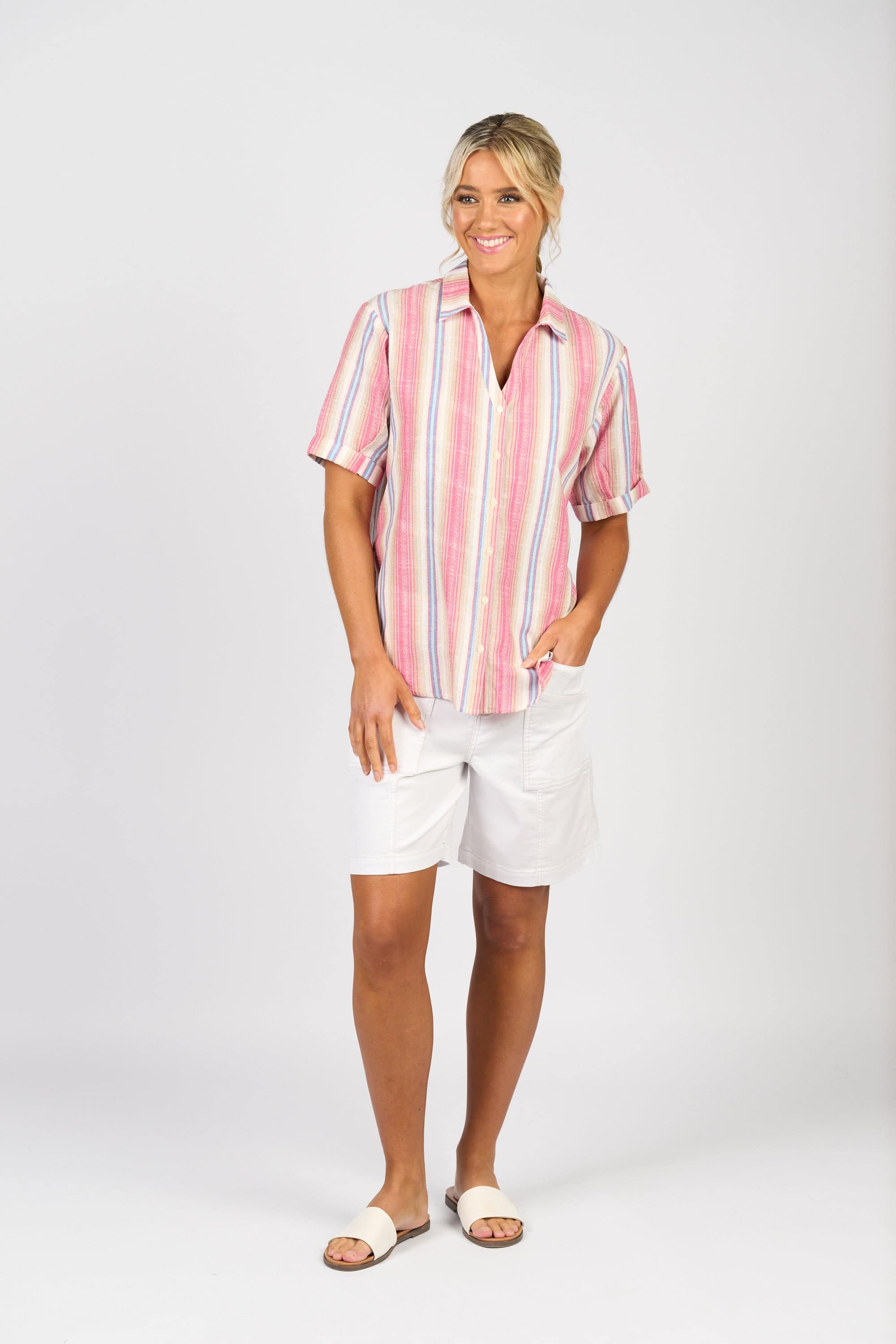 A woman stands smiling against a white background, wearing the Vassalli Shirt - Raspberry Stripe with white shorts and sandals, one hand in her pocket.