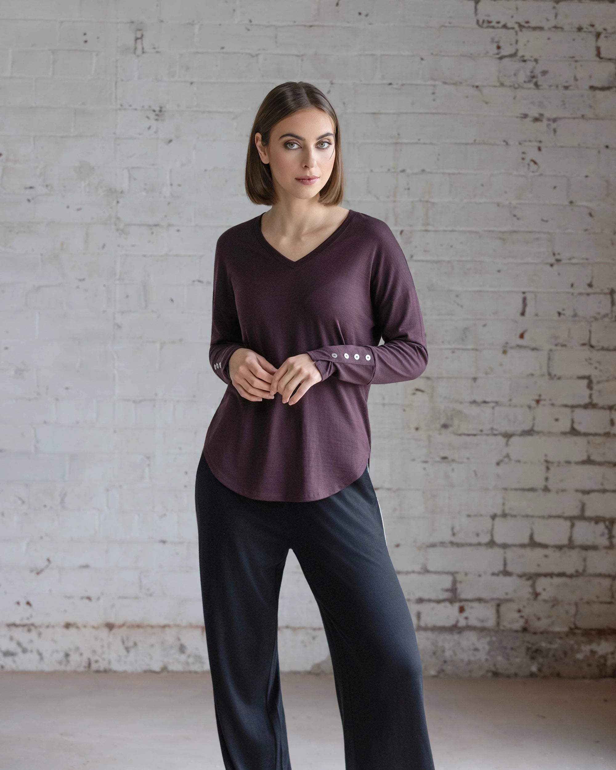 A woman with short brown hair wears the Vassalli Veni Merino Top in Berry, featuring buttoned cuffs, and loose black pants as she stands against a white brick wall, gazing slightly to the side.