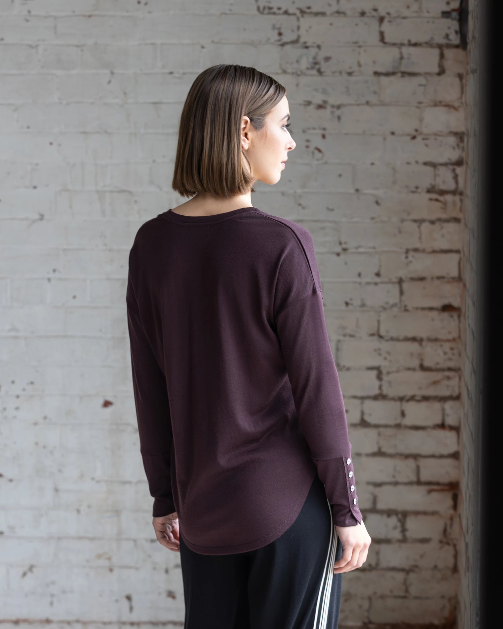 A person with straight, shoulder-length hair wears the Vassalli Veni Merino Top in Berry and black pants with white stripes, standing indoors facing away from the camera against a white brick wall.