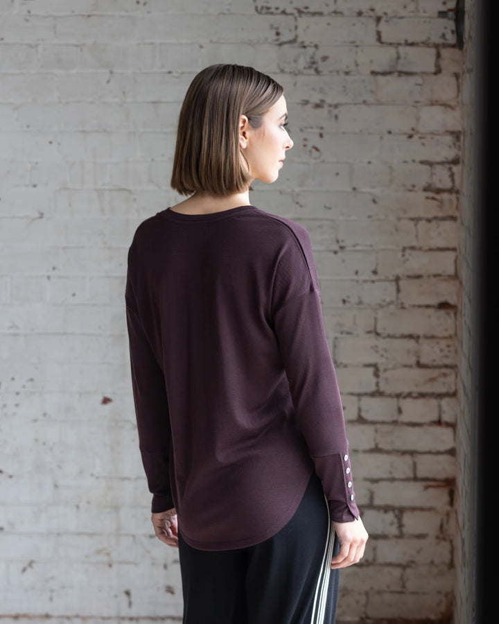 A person with straight, shoulder-length hair wears the Vassalli Veni Merino Top in Berry and black pants with white stripes, standing indoors facing away from the camera against a white brick wall.