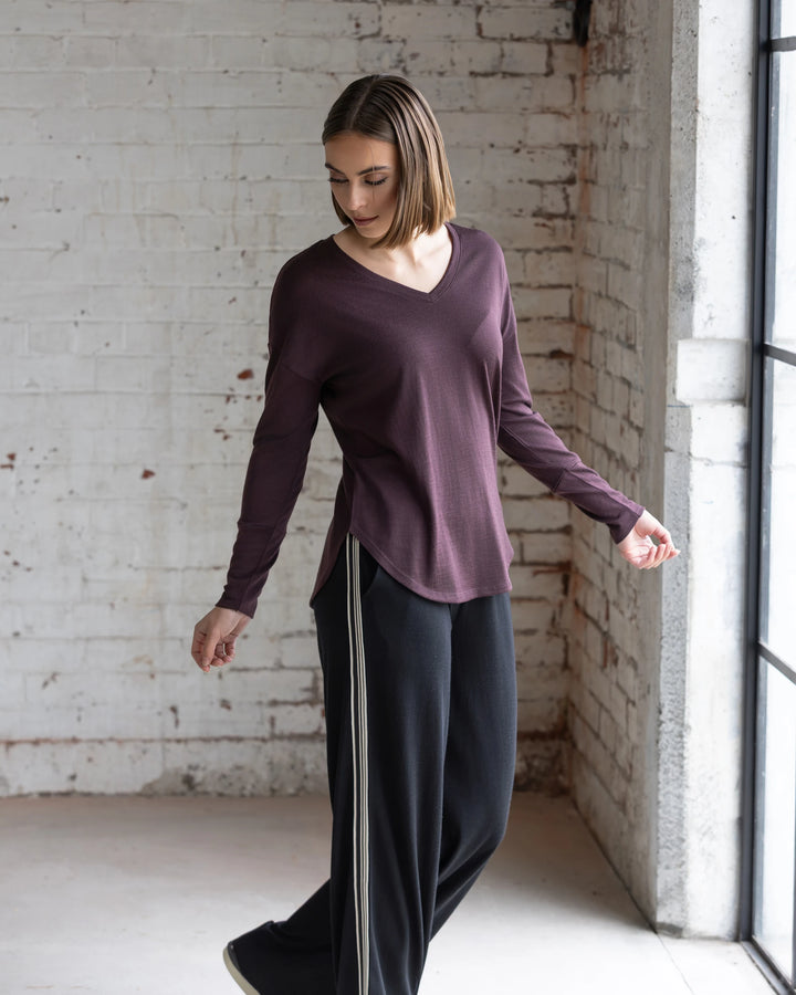 A woman with short brown hair stands by a large window, wearing the Vassalli Veni Merino Top in Berry and black wide-leg pants with white stripes, looking down with one arm slightly extended.