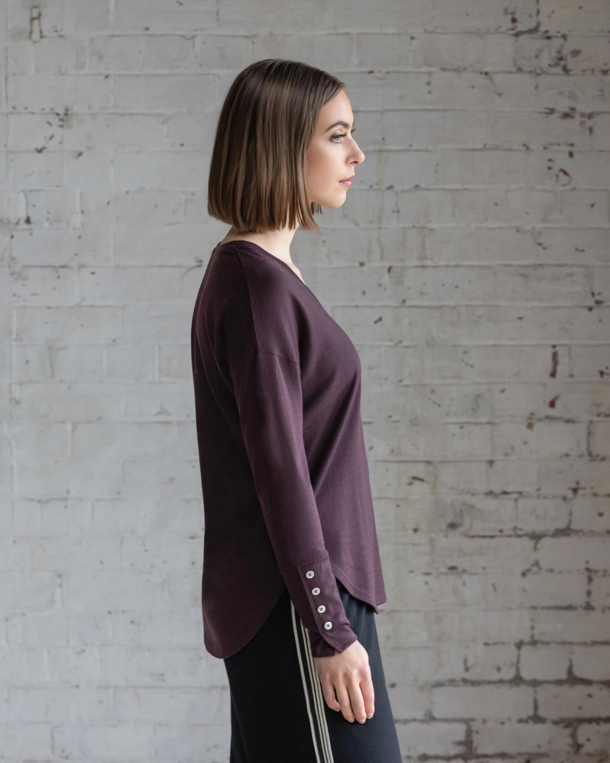 A woman with straight, shoulder-length brown hair stands in profile against a white brick wall, wearing the Vassalli Veni Merino Top in Berry and black pants with side stripes.