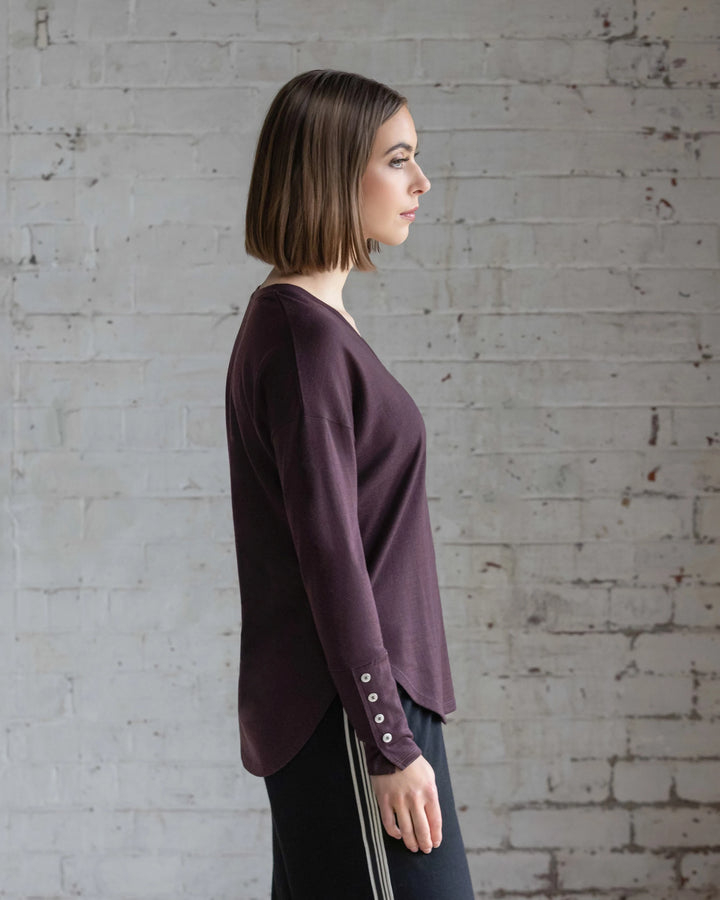 A woman with straight, shoulder-length brown hair stands in profile against a white brick wall, wearing the Vassalli Veni Merino Top in Berry and black pants with side stripes.