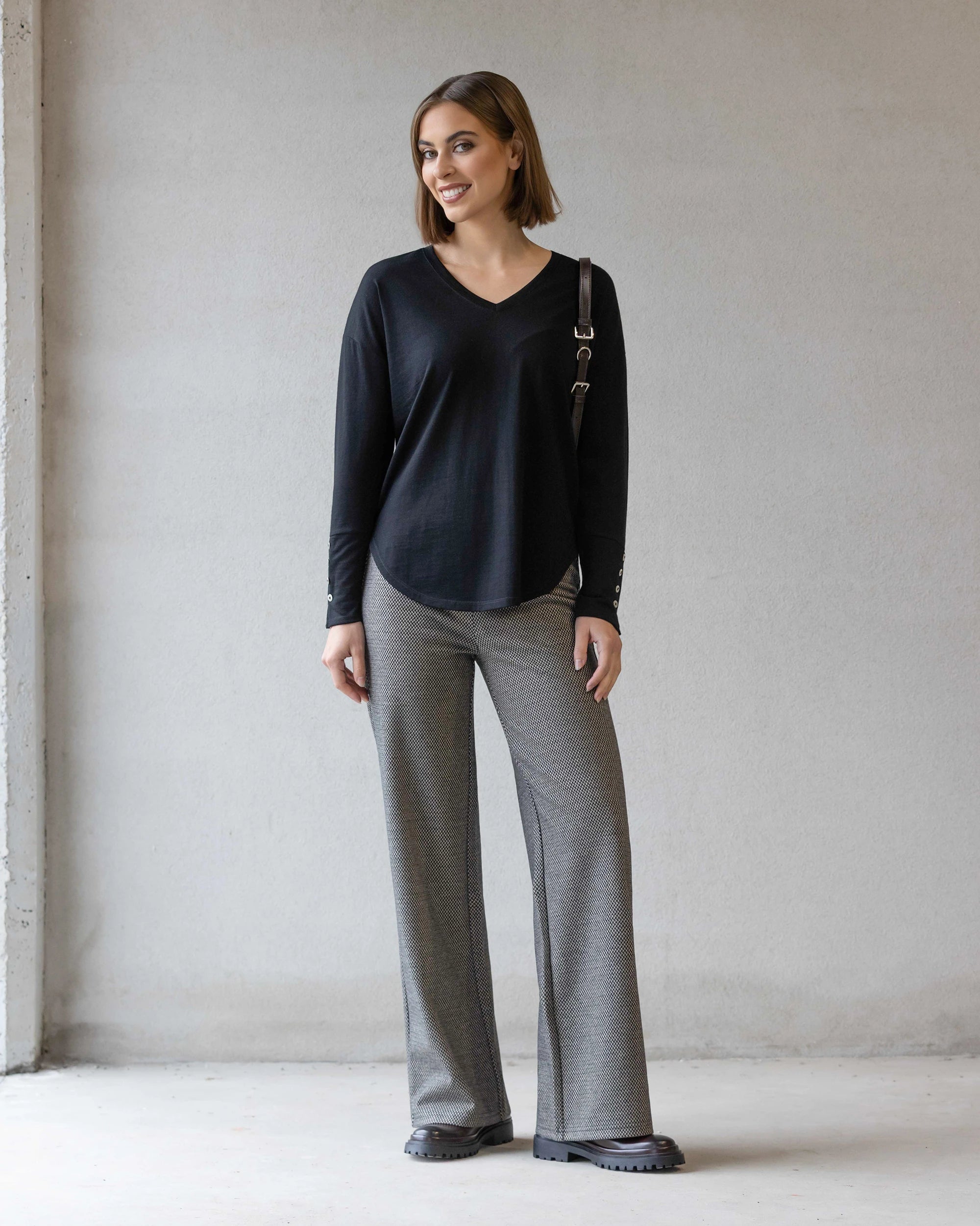 A woman stands indoors against a light-colored wall, smiling. She wears the Vassalli Veni Merino Top in black, gray wide-leg pants, black shoes, and carries a shoulder bag.