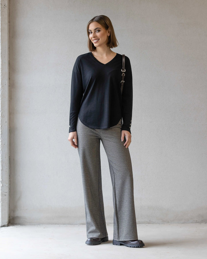 A woman stands indoors against a light-colored wall, smiling. She wears the Vassalli Veni Merino Top in black, gray wide-leg pants, black shoes, and carries a shoulder bag.