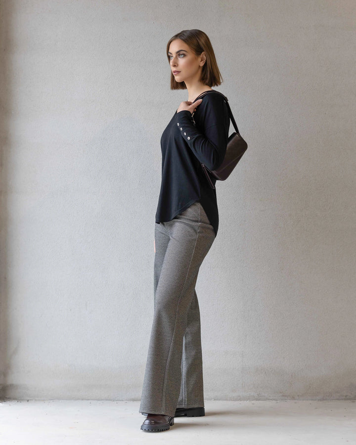 A woman with straight brown hair stands against a plain wall, wearing the Vassalli Veni Merino Top in black, gray flared pants, black shoes, and a black shoulder bag. She faces sideways with a neutral expression.
