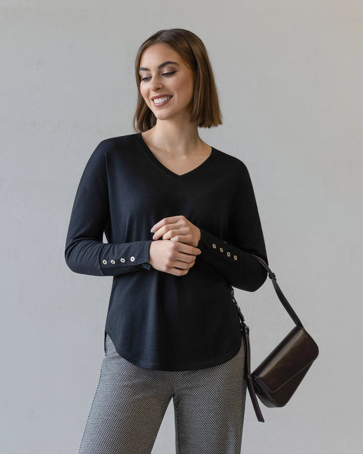 A woman with shoulder-length brown hair wears the Vassalli Veni Merino Top in black, featuring buttoned sleeves, paired with gray patterned pants and a black purse, standing against a plain light background.