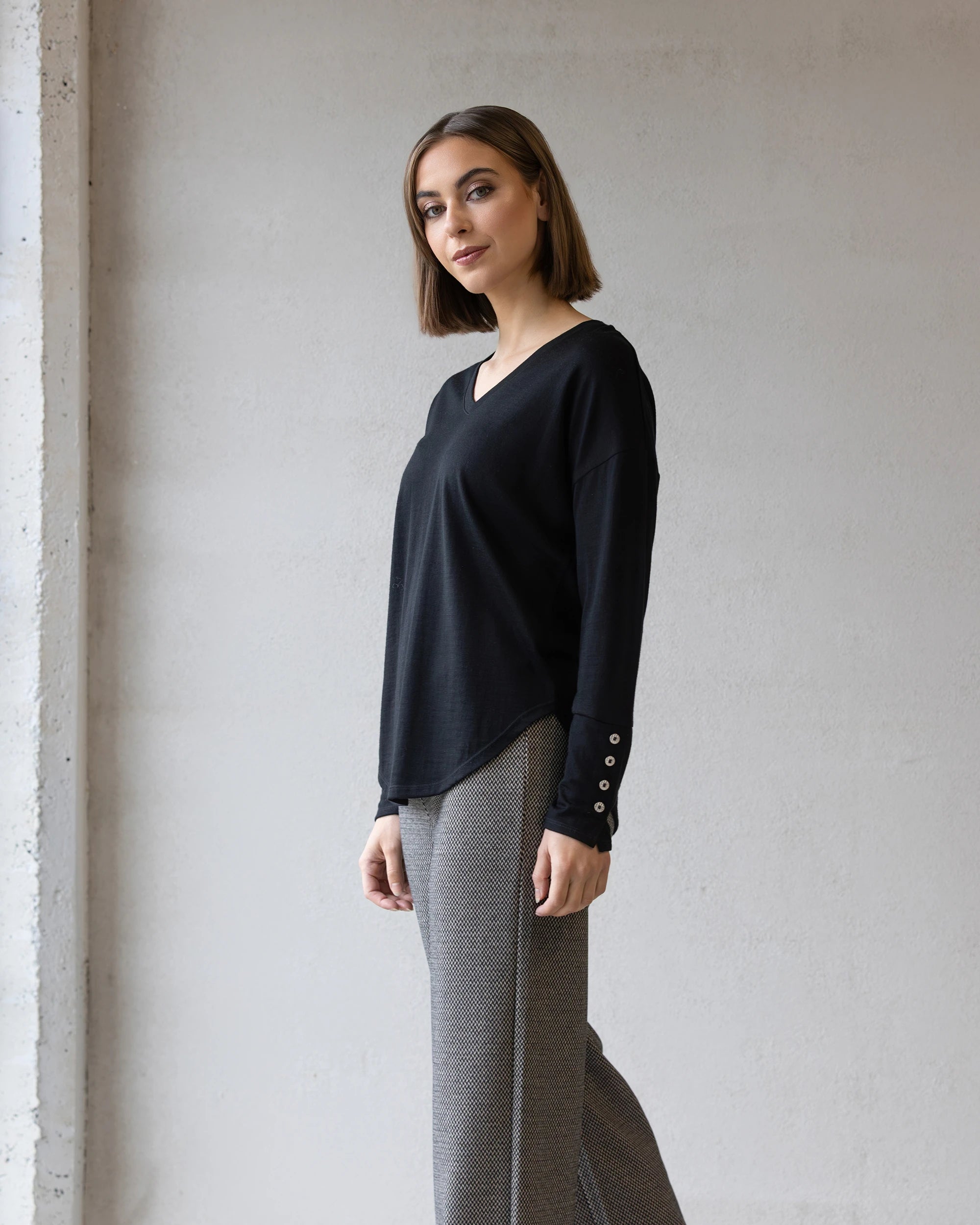 A woman with straight, shoulder-length brown hair stands indoors against a light gray wall, wearing the Vassalli Veni Merino Top in black and gray patterned pants. She faces the camera with a slight smile.