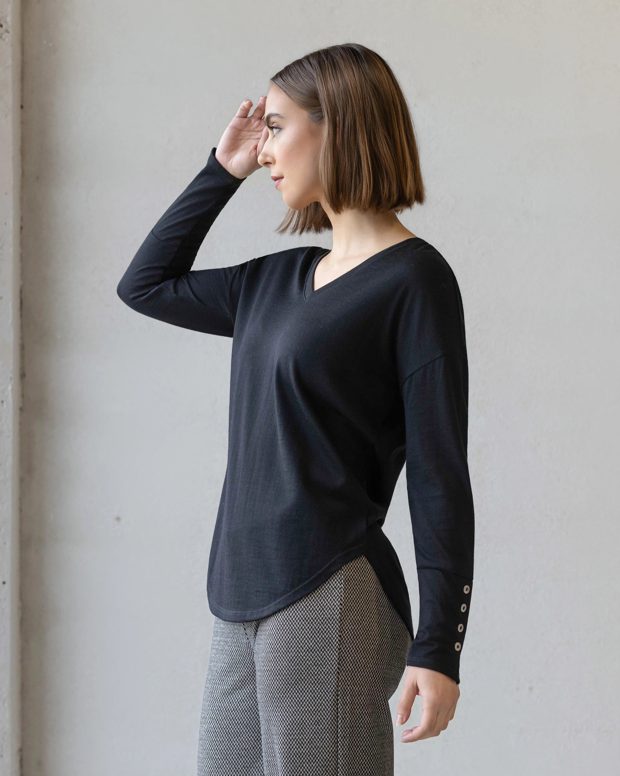 A woman with a straight brown bob stands sideways, looking left with her right hand near her forehead, wearing the Vassalli Veni Merino Top in black and gray patterned pants against a plain light background.