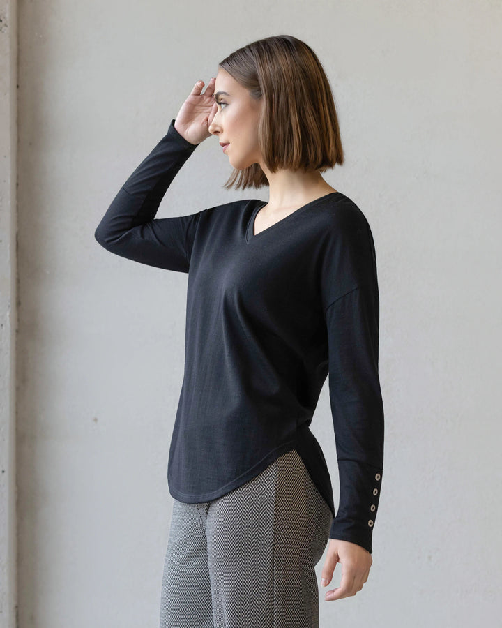 A woman with a straight brown bob stands sideways, looking left with her right hand near her forehead, wearing the Vassalli Veni Merino Top in black and gray patterned pants against a plain light background.