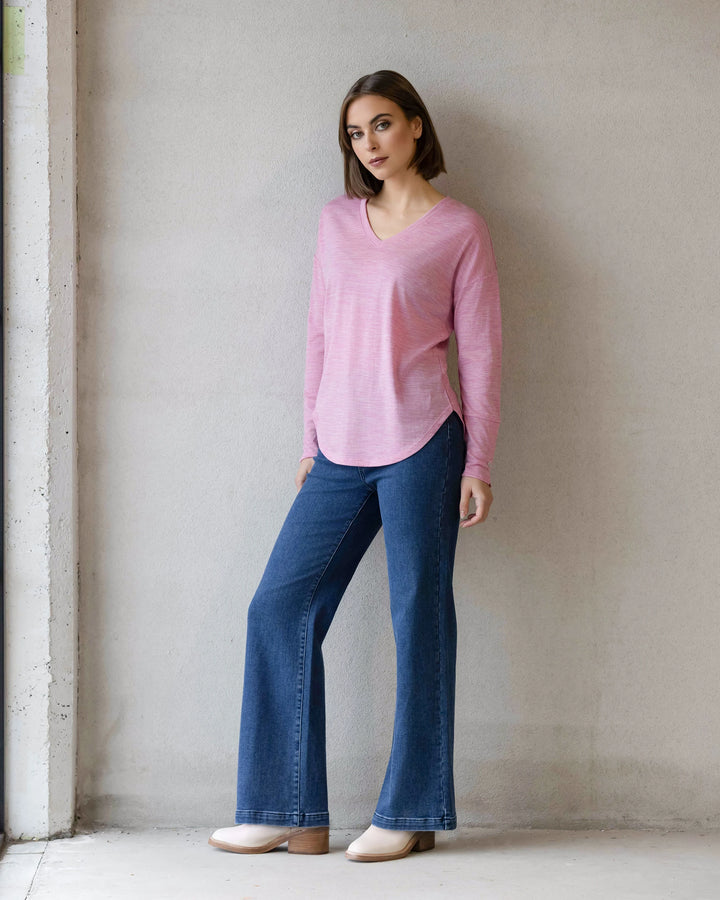 A woman with straight brown hair and a neutral expression stands by a light wall, wearing the Vassalli Veni Merino Top in Pink Marle, blue flared jeans, and light shoes—a perfect winter layering style.