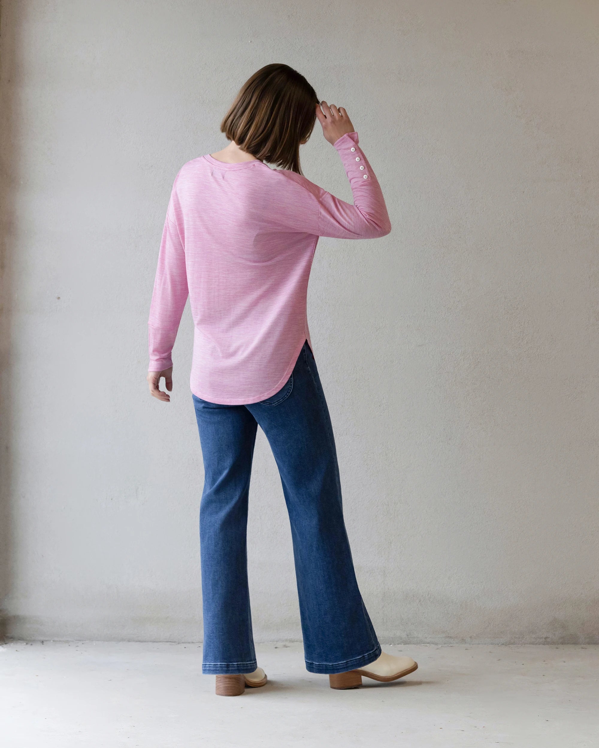 A person with short brown hair stands facing a light wall, wearing the Vassalli Veni Merino Top in Pink Marle, blue jeans, and beige shoes, with their back to the camera and one hand raised to their head.