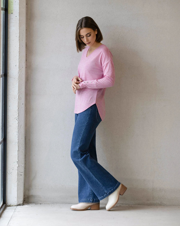 A woman with a short brown bob wears the Vassalli Veni Merino Top in Pink Marle, paired with blue wide-leg jeans and beige shoes. She stands indoors by a window, hands loosely clasped—ideal winter layering.