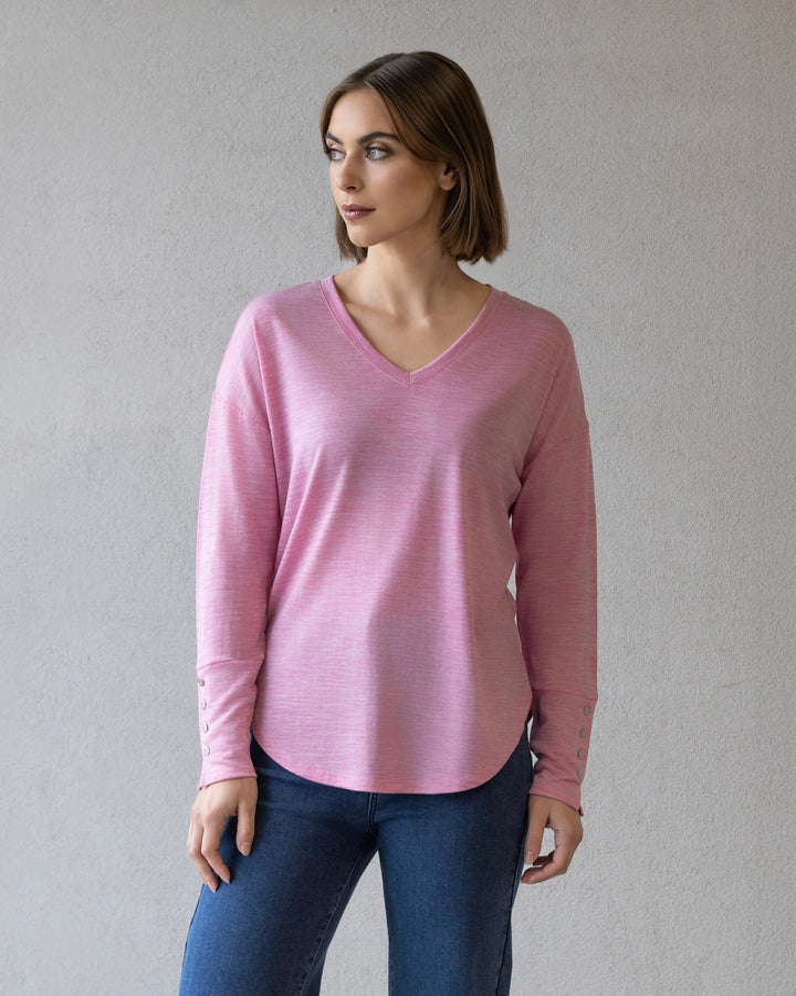 A person with short brown hair wears the Vassalli Veni Merino Top in Pink Marle and blue jeans, perfect for winter layering, standing against a plain light gray wall.