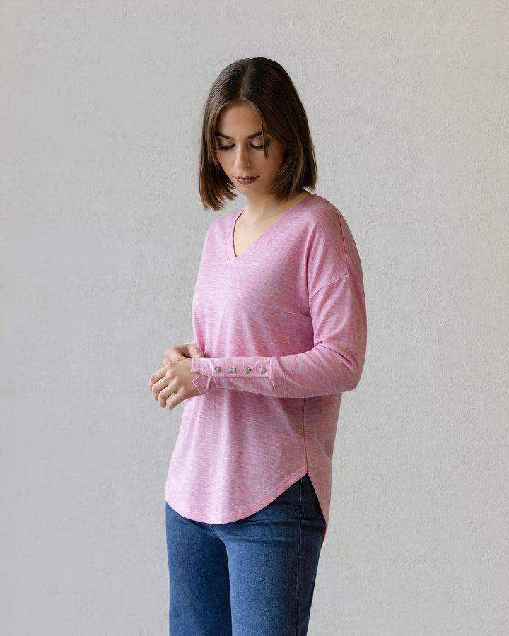 A woman with shoulder-length brown hair wears the Vassalli Veni Merino Top in Pink Marle and blue jeans, standing against a plain light background while looking down and touching her hands—perfect for winter layering.
