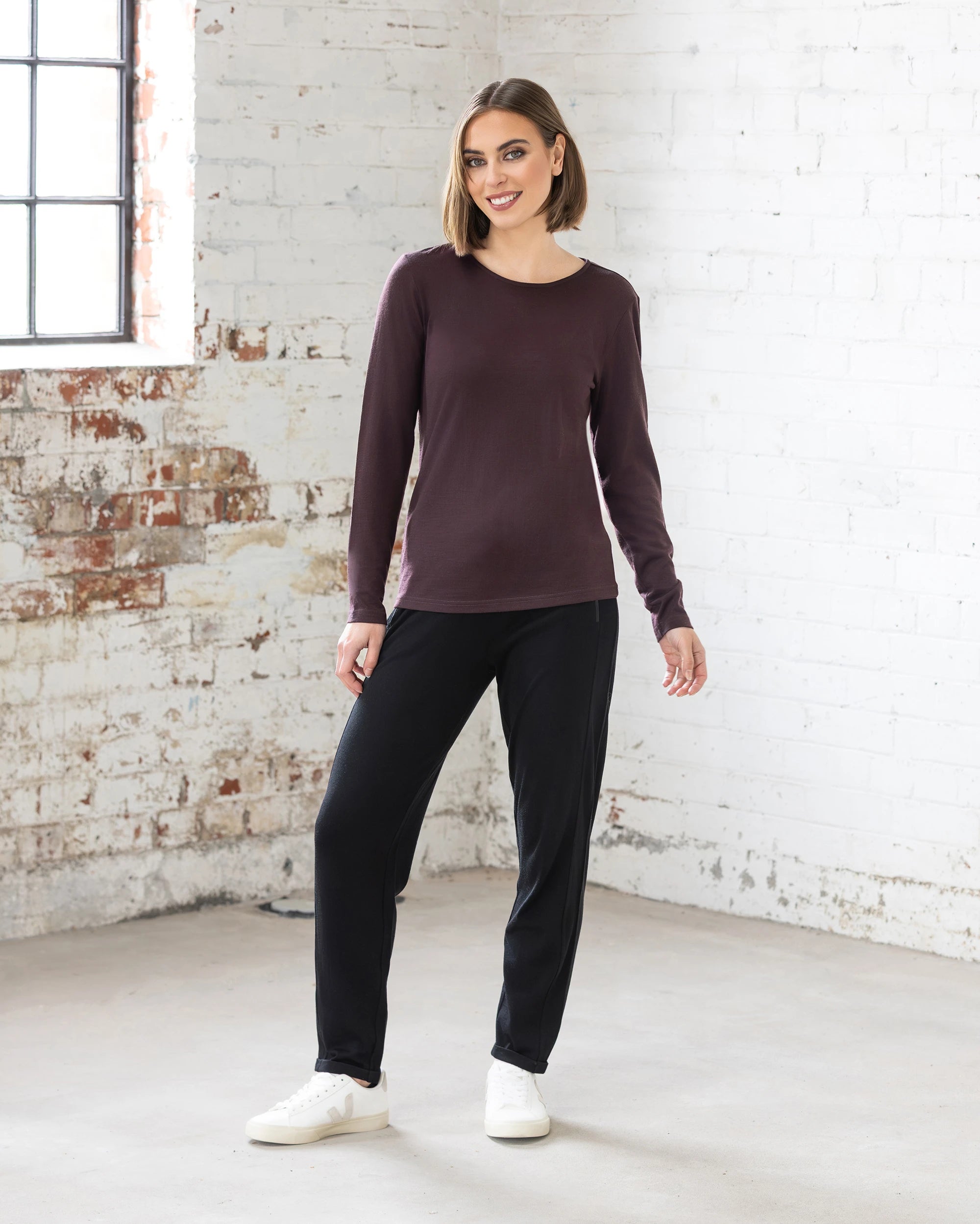 A woman with a bob stands indoors against a white brick wall, smiling in the Vassalli Tori Merino Crew - Berry, paired with black pants and white sneakers. She stands casually with one hand relaxed at her side.
