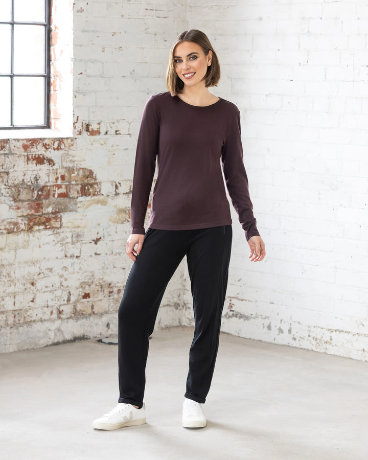 A woman with a bob stands indoors against a white brick wall, smiling in the Vassalli Tori Merino Crew - Berry, paired with black pants and white sneakers. She stands casually with one hand relaxed at her side.