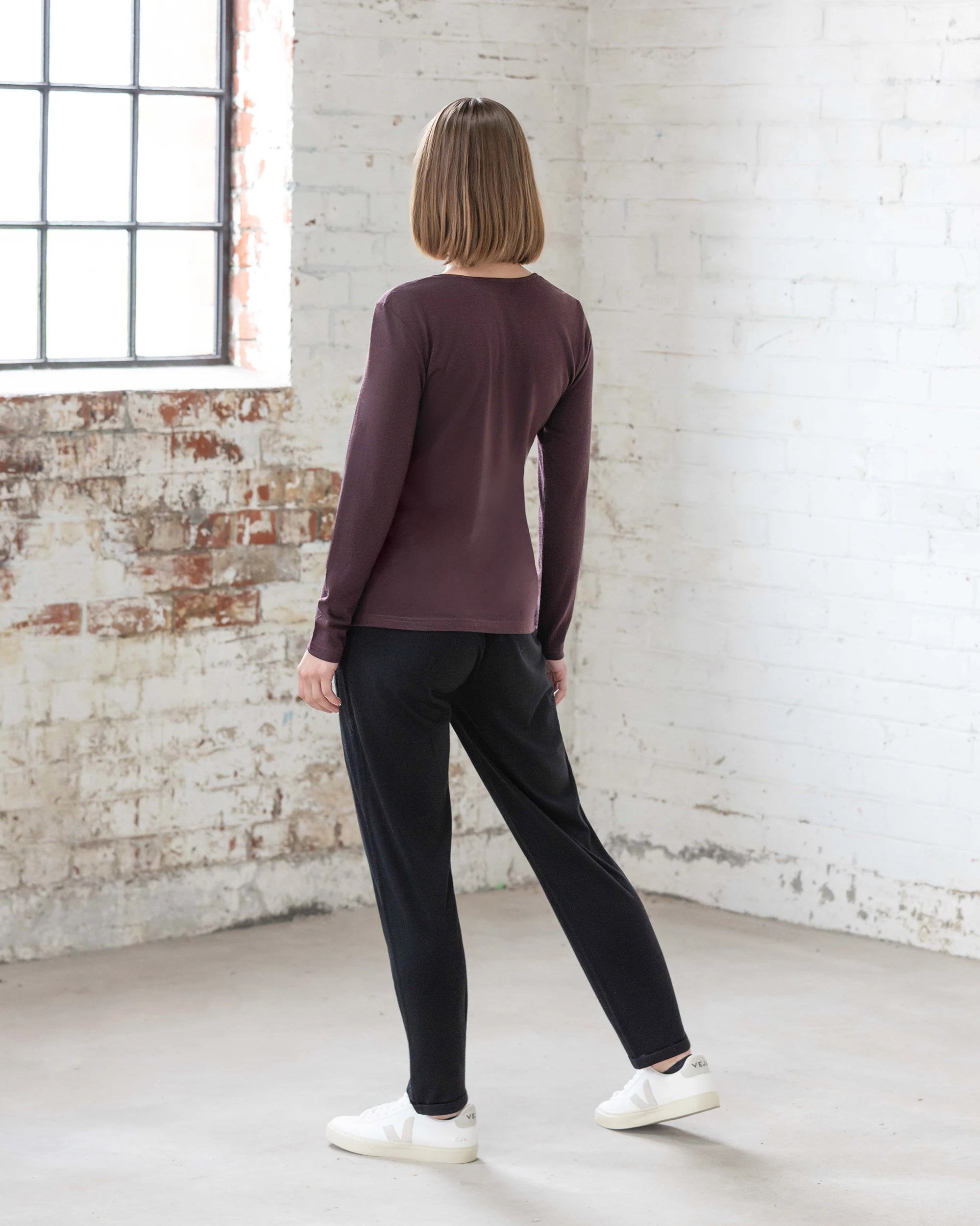 A person with short, light brown hair stands indoors by a white brick wall, wearing the Vassalli Tori Merino Crew in Berry as a versatile layer, styled with black pants and white sneakers. Natural light filters in from the left window.
