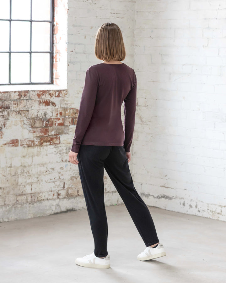 A person with short, light brown hair stands indoors by a white brick wall, wearing the Vassalli Tori Merino Crew in Berry as a versatile layer, styled with black pants and white sneakers. Natural light filters in from the left window.