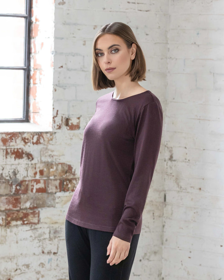 A woman with a bob haircut stands indoors near a white brick wall and window, wearing the Vassalli Tori Merino Crew - Berry—an ideal layering essential—paired with black pants.
