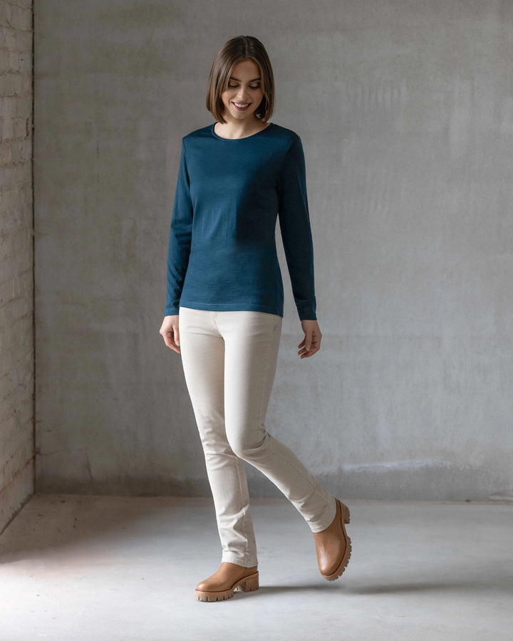 A woman wearing the Vassalli Tori Merino Crew - Dragonfly, paired with light beige pants and brown shoes, stands in a minimalistic room with gray walls and a concrete floor, looking down and smiling.