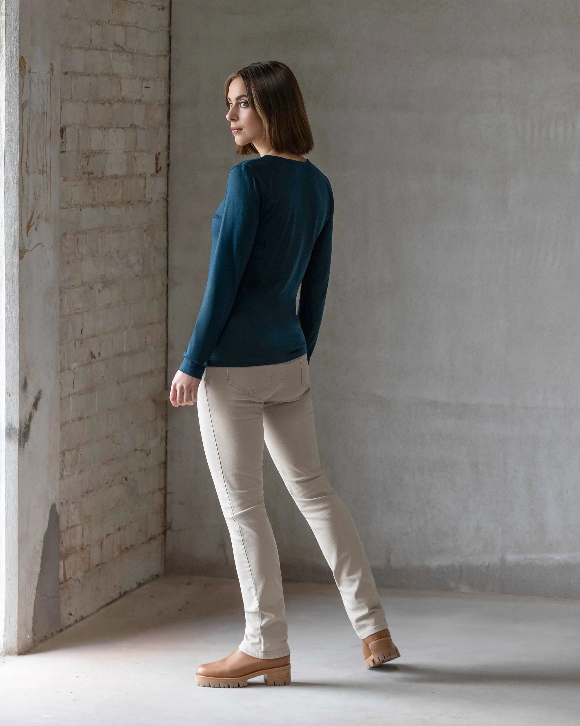 A woman with straight brown hair wears the Vassalli Tori Merino Crew in Dragonfly, paired with beige pants and tan boots, as she stands indoors on a concrete floor, glancing over her shoulder towards the window.