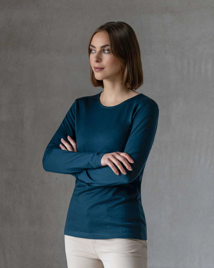A woman with straight, shoulder-length brown hair stands against a gray textured background wearing the Vassalli Tori Merino Crew in Dragonfly and light pants, arms crossed and a neutral expression—perfect for layering.