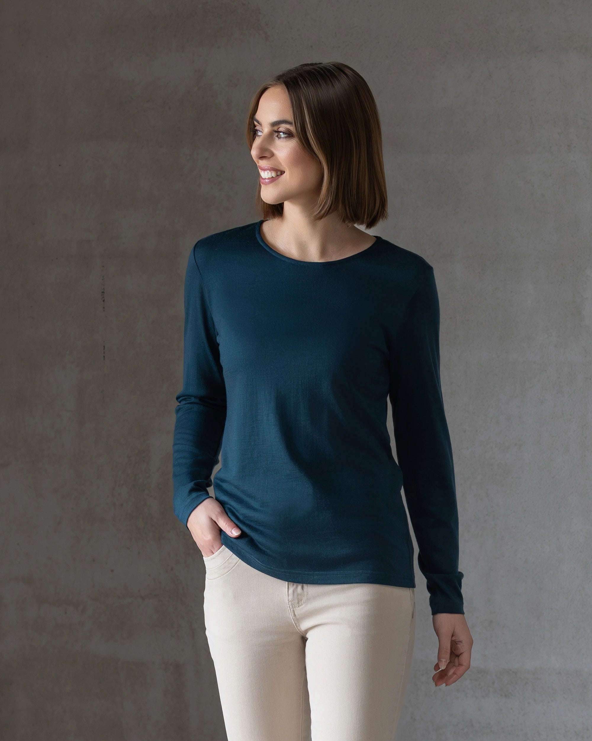 A woman with straight brown hair wears the Vassalli Tori Merino Crew in Dragonfly and beige pants, standing against a gray textured wall, looking to her left and smiling with one hand in her pocket.