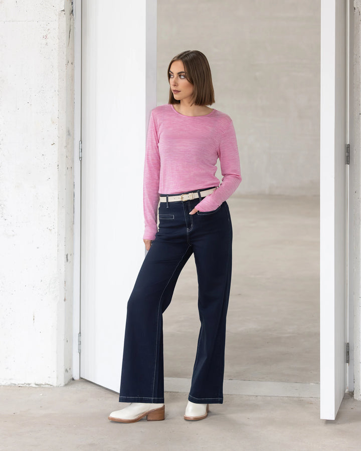 A woman with a bob haircut wears the Vassalli Tori Merino Crew in Pink Marle, paired with dark wide-leg jeans, a beige belt, and white boots. She stands casually in a minimalist white doorway—perfect for winter layering.