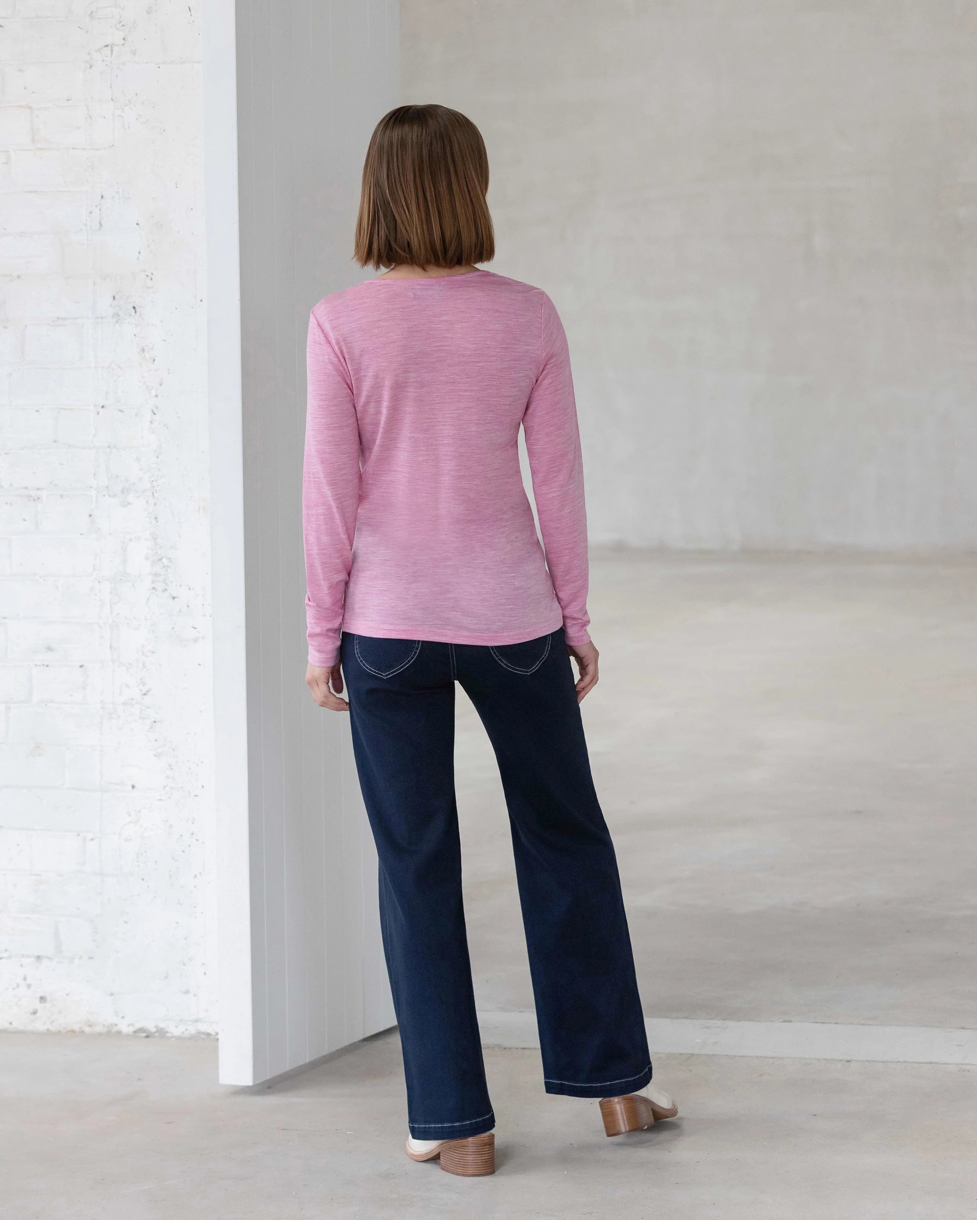 A person with straight, shoulder-length brown hair stands indoors wearing the Vassalli Tori Merino Crew in Pink Marle, paired with dark blue jeans and light shoes. The minimal white-walled setting makes it ideal for winter layering.
