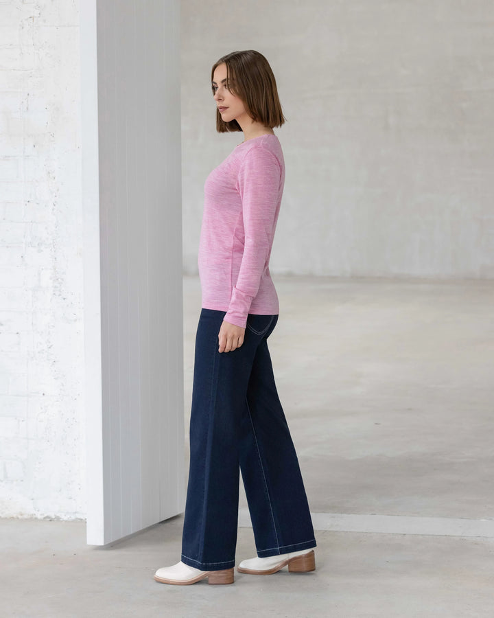 A woman with straight brown hair stands in profile, wearing the Vassalli Tori Merino Crew in Pink Marle, dark blue wide-leg jeans, and white shoes in a minimalist, light-toned room—ideal for winter layering.