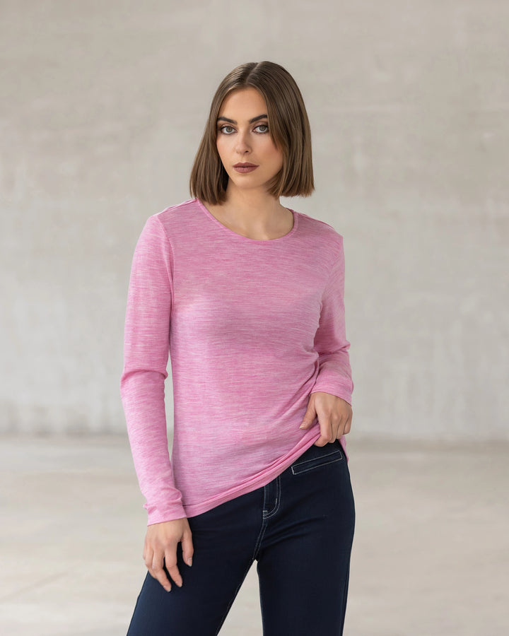 A woman with straight brown hair wears the Vassalli Tori Merino Crew in Pink Marle, styled with dark jeans against a light indoor background—an ideal winter layering look.