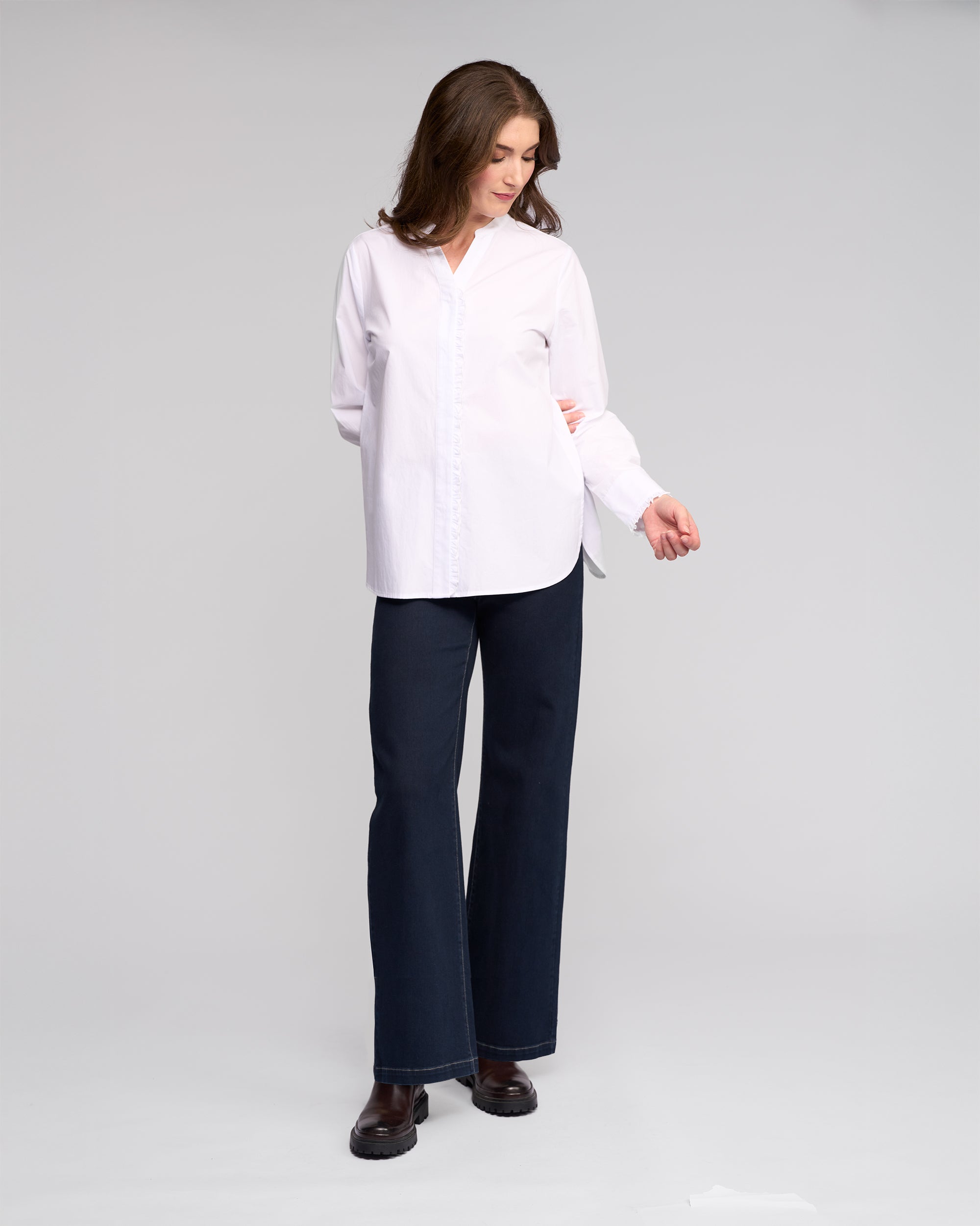 A woman with long brown hair wears the Vassalli Manhattan Shirt in white, paired with dark wide-leg jeans and black shoes. She stands against a light grey background, looking down to her left with her hands behind her back.