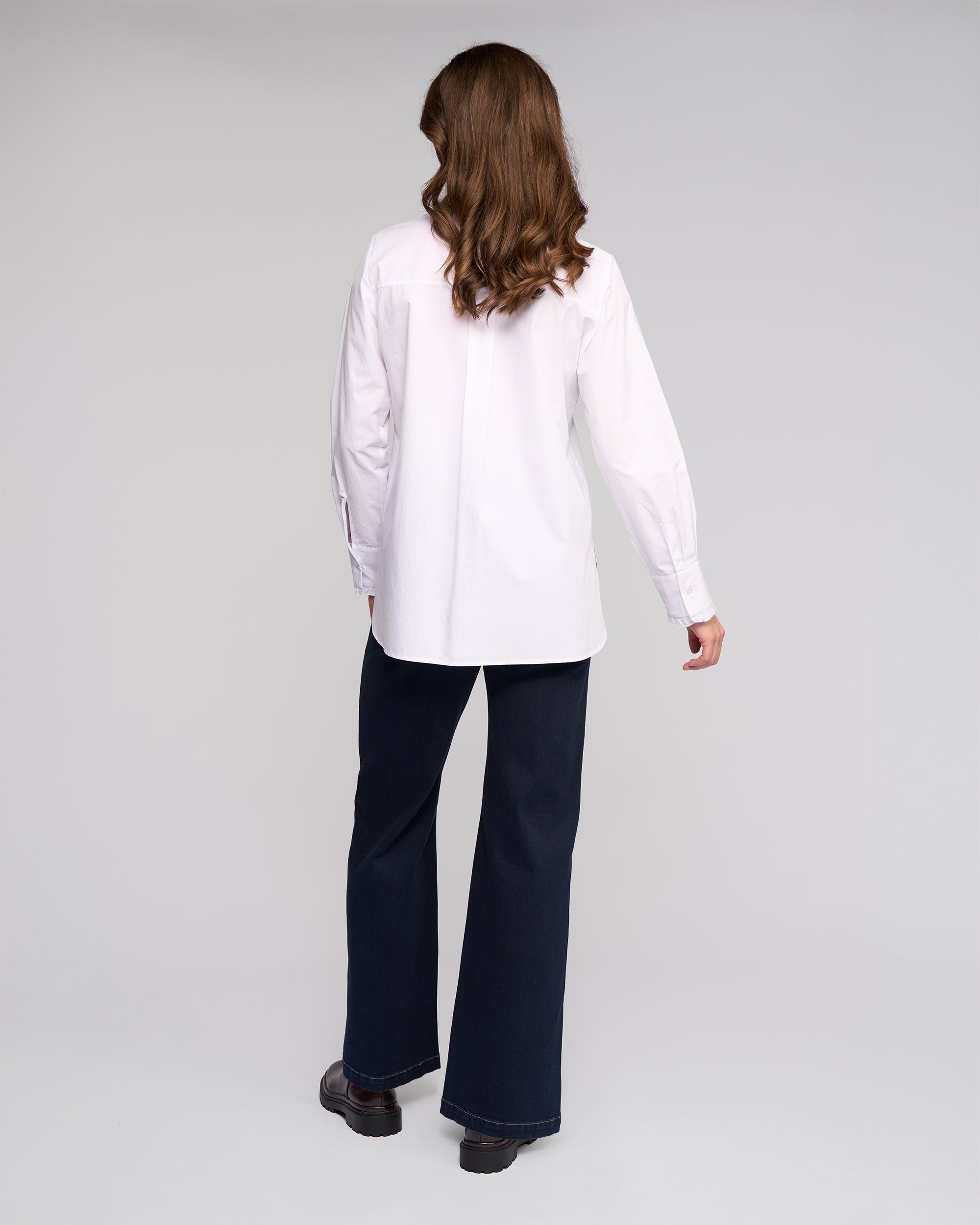 A woman faces away, wearing the Vassalli Manhattan Shirt in white with long sleeves, paired with dark flared pants and black shoes, set against a plain light gray background.