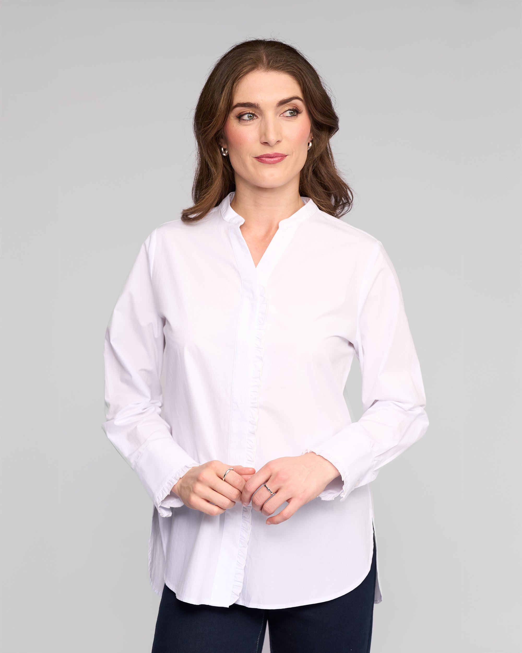 A woman with long brown hair wears the Vassalli Manhattan Shirt in white with dark pants, standing against a plain light gray background. She looks slightly left with a neutral expression and her hands clasped in front of her.