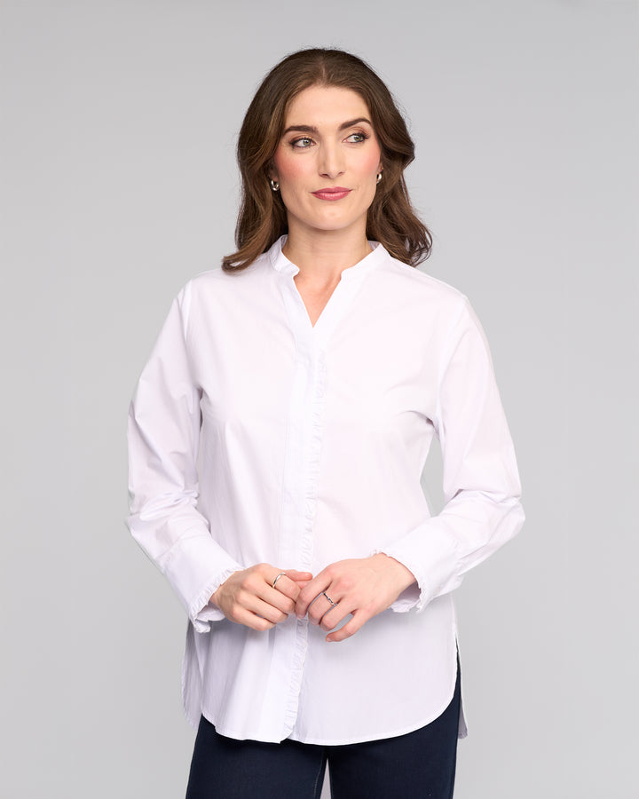 A woman with long brown hair wears the Vassalli Manhattan Shirt in white with dark pants, standing against a plain light gray background. She looks slightly left with a neutral expression and her hands clasped in front of her.