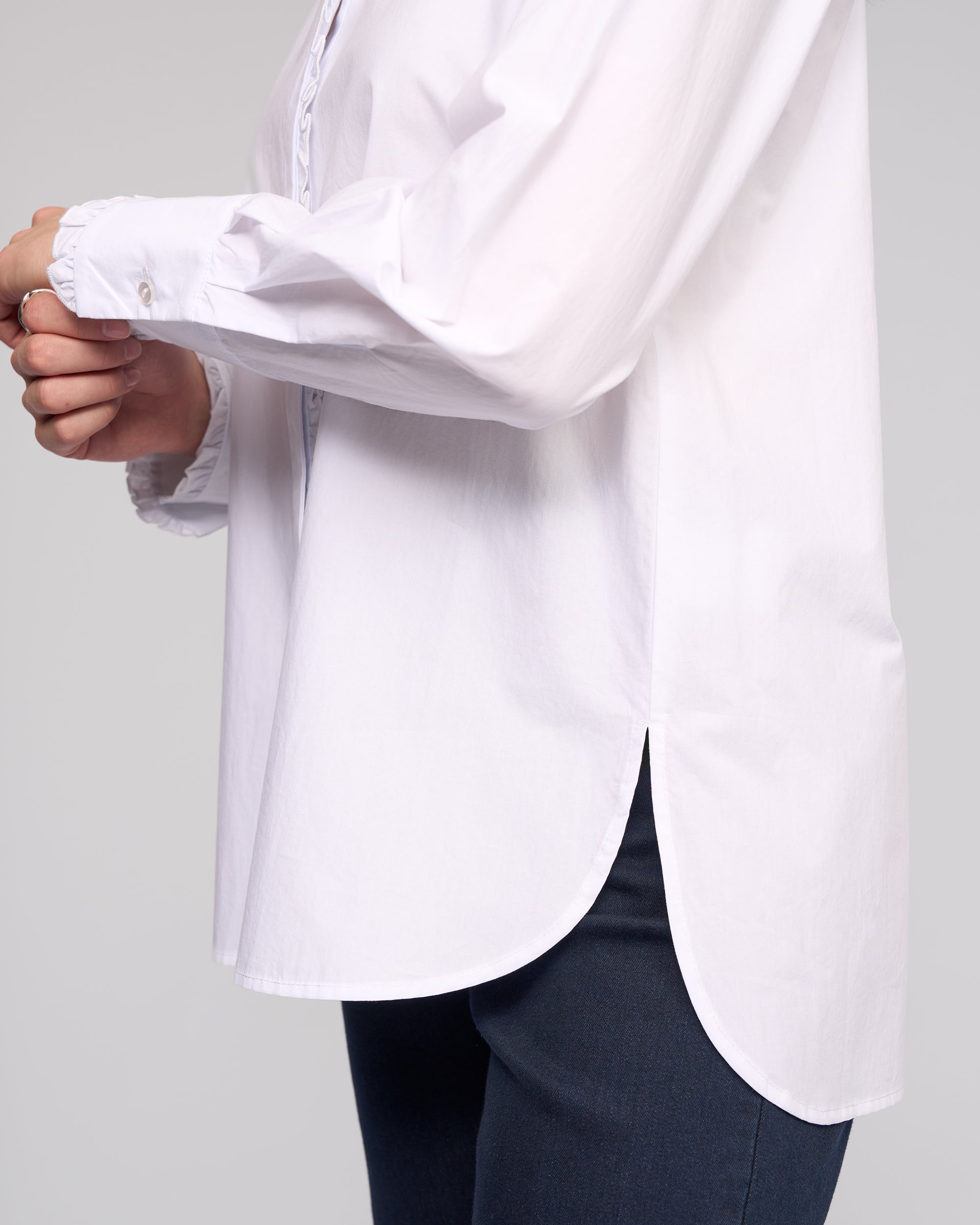 A person wearing the Vassalli Manhattan Shirt - White with a rounded hem and dark pants, shown in profile and cropped at the shoulders and upper legs.